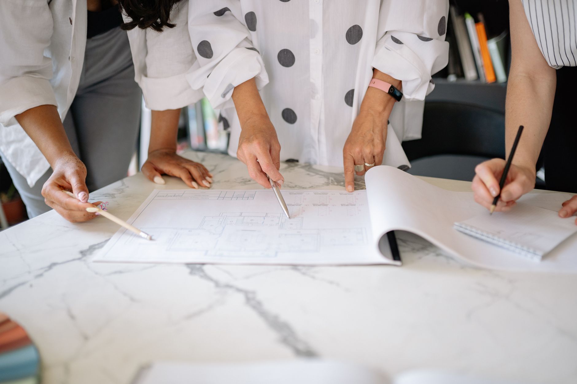 Three people lean over a marble table covered with floor plans, pointing at details and sketching in a notebook.