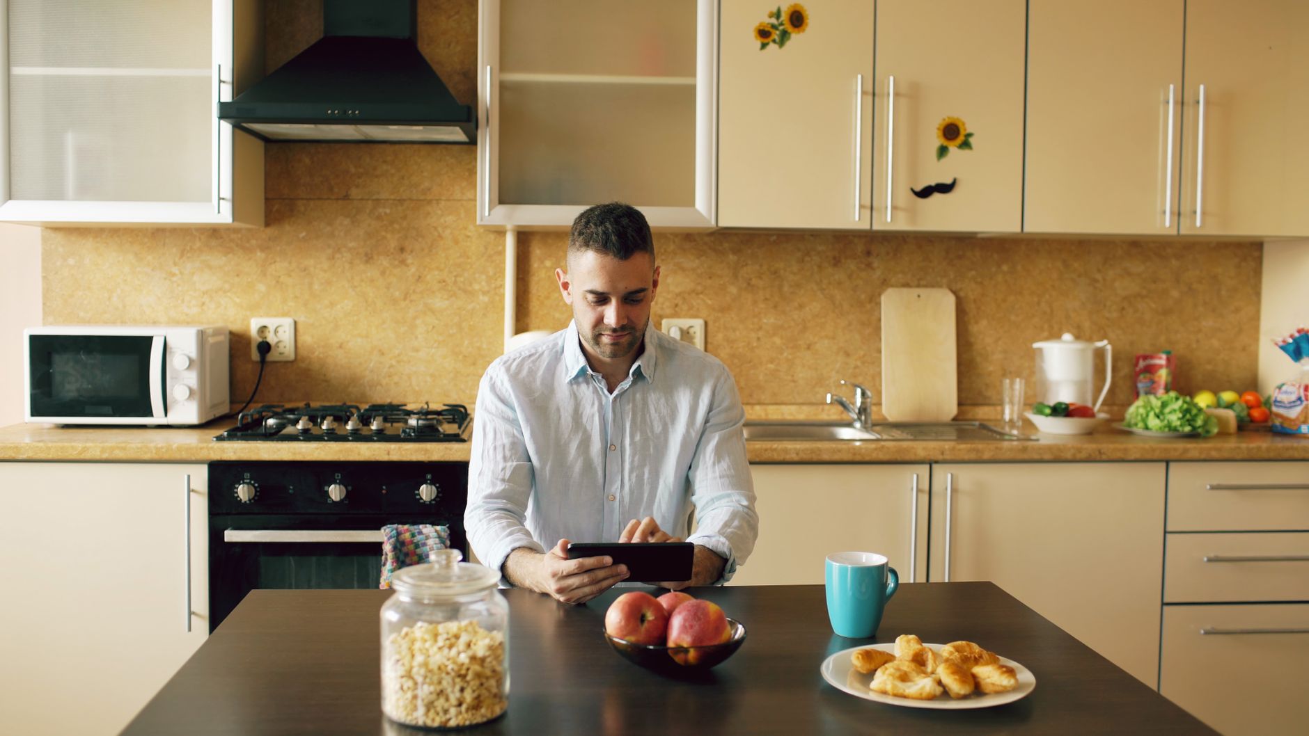 A man at a kitchen table reading on a tablet, with a blue mug, a bowl of apples and a plate of pastries in front of him.