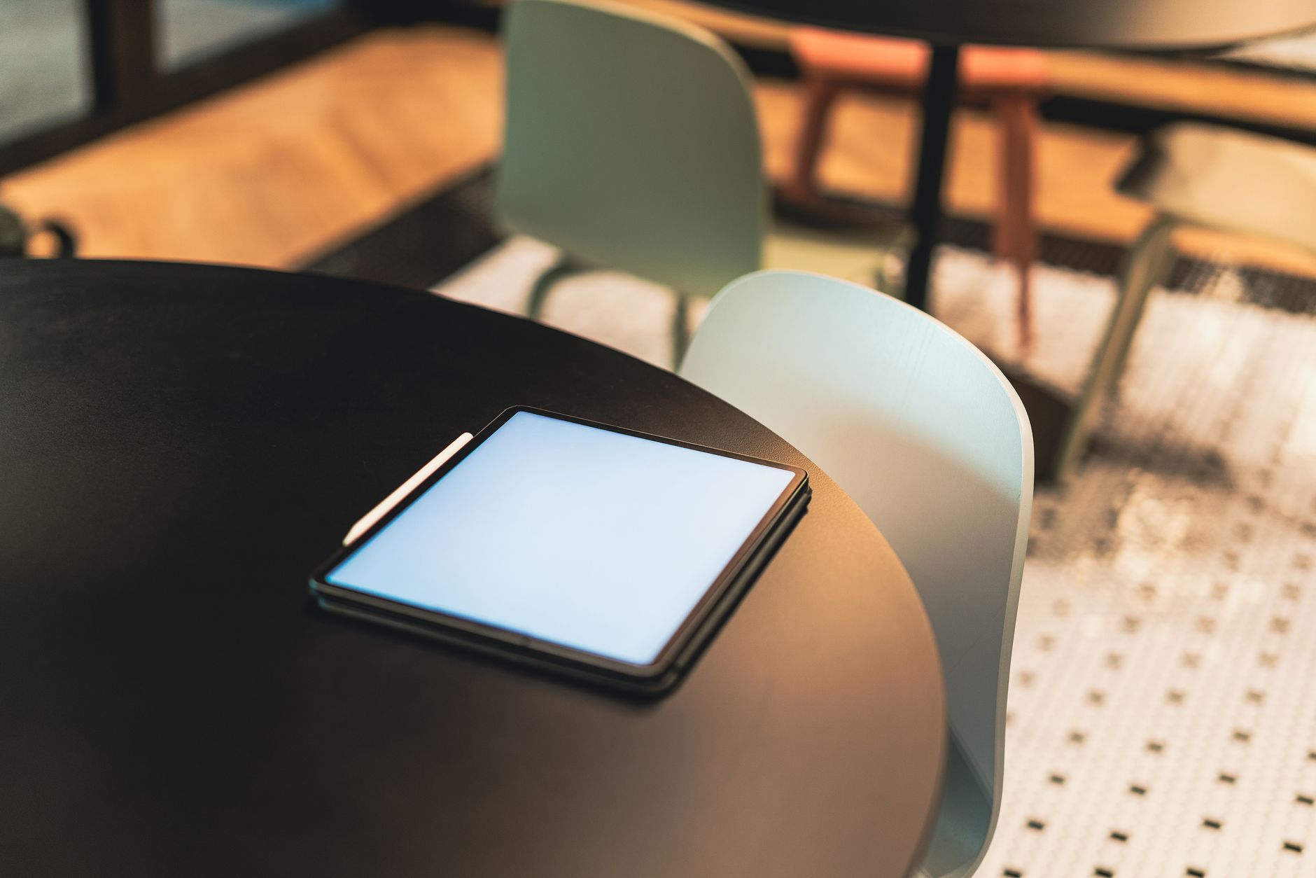 A tablet with a bright screen resting on a dark round table in a softly lit cafe, with pale chairs behind it.