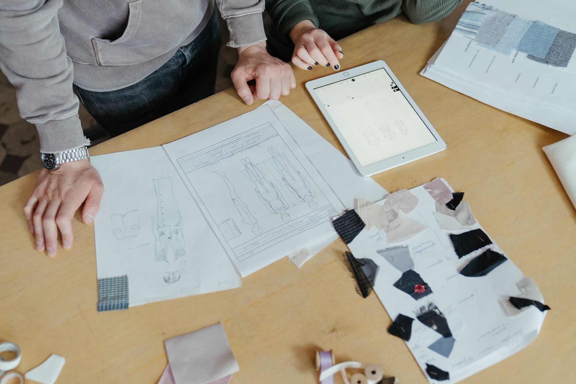 Two people at a wooden worktable with design sketches, fabric samples and a tablet, planning a project together.