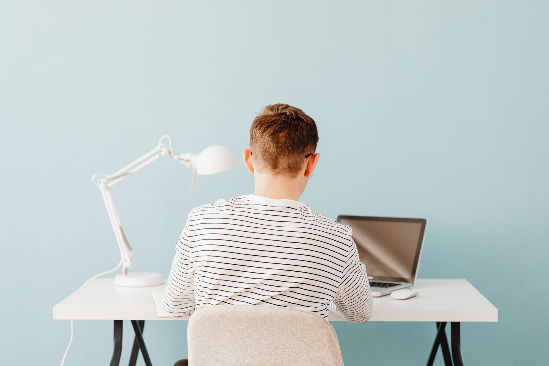 Teenager using laptop at desk under lamp in a minimalist home setting.